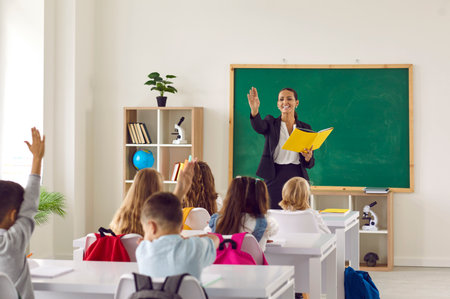 Friendly female teacher conducts lesson for children in modern elementary school. Children sit at...