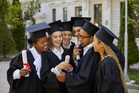 Happy smiling graduate multiracial people in graduation gowns and caps looking at diploma after...