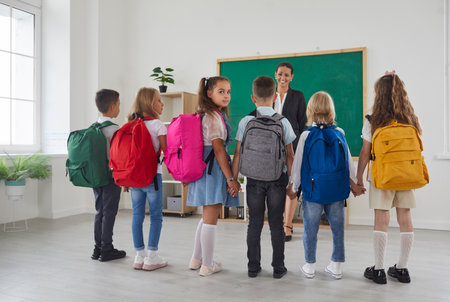 Schoolchildren with colorful backpacks on their shoulders are holding hands while talking to teacher...