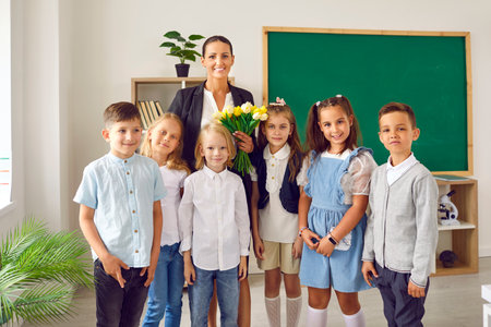 Cute happy little children boys and girls posing for group portrait together with woman teacher holding flowers in hand standing in classroom with green board for elementary school. Pre-teens, kidsの写真素材