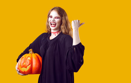 Funny, happy, cheerful woman in spooky costume standing isolated on yellow background, holding orange pumpkin, smiling and showing something on copyspace sideの写真素材