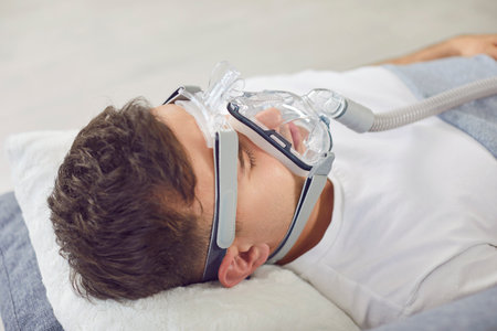 Man lying on the couch at home, breathing through an oxygen mask. He is dealing with asthma, apnea...