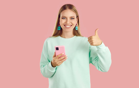 Happy blondy girl in mint sweatshirt and earrings is showing thumb up holding smartphone on pink...