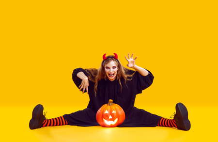 Frightening woman with Halloween makeup sitting with pumpkin on orange background. Woman with mad...