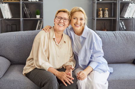 Cheerful adult woman and her elderly mother posing together on sofa, embracing warmly and smiling...