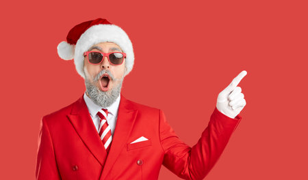 Funny man in Christmas costume pointing aside. Happy surprised man in suit, tie, Santa cap, white gloves and sunglasses standing on red studio background and pointing index finger to sideの写真素材