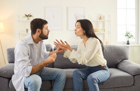 Angry frustrated husband and wife sitting on sofa in bright living room and arguing loudly. Unhappy family couple shouting and gesturing emotionally, experiencing marriage problems during conflict.の写真素材