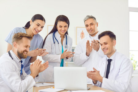 Happy smiling doctors rejoicing at success in treatment applauding standing at desk in clinic during meeting looking at laptop. Successful team of young medical staff in hospital. Health care conceptの写真素材