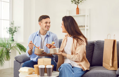 Young married couple eating meal from takeaway containers while talking on the sofa at home. Casual and intimate moment, highlighting the convenience and comfort of food delivery for a relaxed lunch.の写真素材