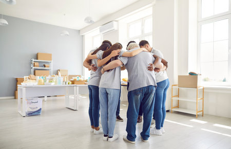 Group of volunteers of different ages indoors standing in a circle hugging, view from the back. Employees of a charity fund before starting work. Concept homeless center, humanitarian projectの写真素材