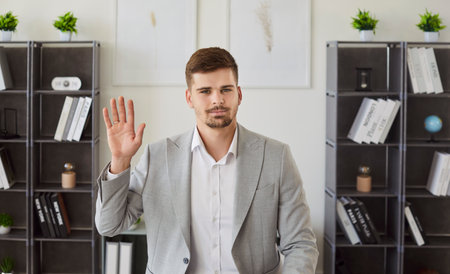 Young man waving hand looking at camera having video call on workplace in modern office. Male employee chatting online during business conference call. Internet communication and technology concept.の写真素材