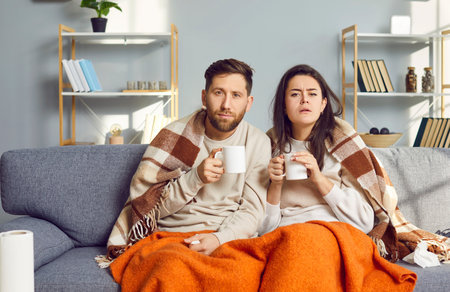Couple relaxing on couch under blanket at home in living room. Sleepy spouses wrapped in warm blankets sitting close to each other, drinking hot tea or coffee and watching at somethingの写真素材