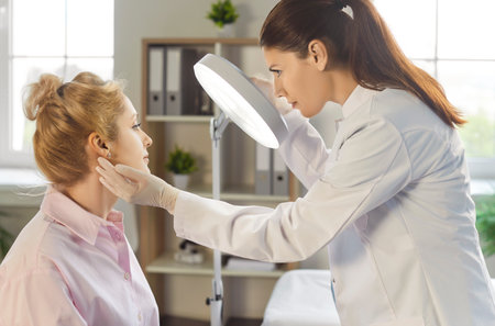 Doctor beautician, cosmetologist examining female patient face, dermatologist nurse wearing sterile gloves under magnifying lamp in clinic room, diagnosing and treating consultation medical careの写真素材