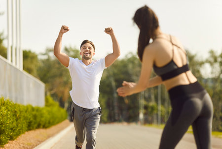 Happy athletic man running toward young woman with arms raised in victory. Joyful guy beating record, celebrating fitness achievement while training outdoors with supportive partner in summer park.の写真素材