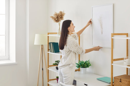 Cheerful young woman arranging piece of art on wall, hanging picture on wall in her sunny, contemporary apartment, smiling and organizing, decorating her living space. Housing improvement conceptの写真素材