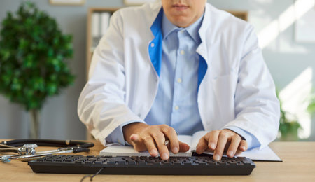 Cropped image of unknown doctor sitting at workplace in his cabinet, typing on keyboard, working with computer, doing electronic medical record, posing in medical clinic. Medicine conceptの写真素材