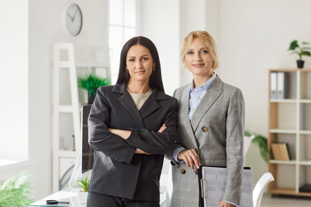 Two positive businesswomen office workers women together cooperating on project, standing in modern room looking at camera, smiling, mentor teaching intern, female colleagues in friendly relationsの写真素材