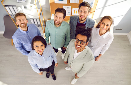 Team of happy diverse business people at work. High angle shot of a group of different young mixed race men and women standing in the office, looking up at the camera, and smilingの写真素材