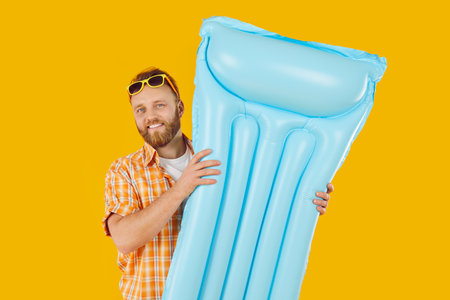 Funny young smiling man wearing beach clothes in sunglasses holding inflatable mattress isolated on...