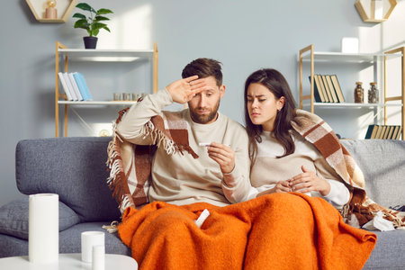 Sick couple covered in blankets sitting on sofa at home with seasonal flu or cold. Woman looking at thermometer of man checking temperature suffering from headache feeling unhealthy with influenza.の写真素材