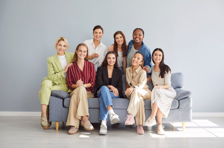 Diverse group of smiling women sitting on comfortable sofa in bright modern room. Females looking at camera, making group photo against light gray background, creating sense of unity and support.の写真素材