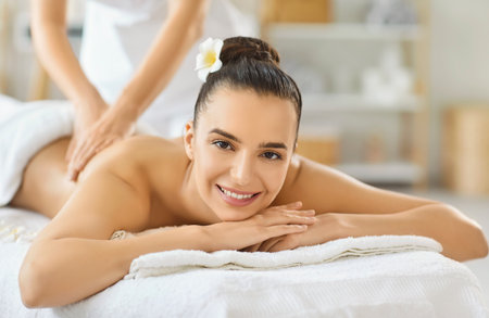 Portrait of smiling young woman lying on massage table and enjoying relaxing back massage in spa salon. Happy female client receiving wellness therapy from professional masseur in beauty center.の写真素材