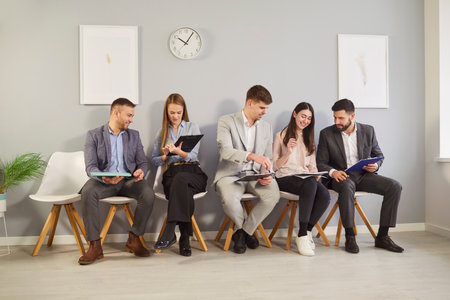 Group of business people sitting on chairs with clipboards, chatting and checking papers. Smiling...