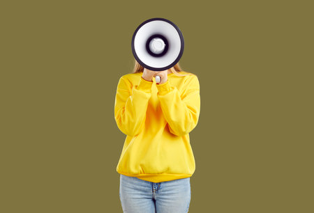 Young woman makes loud announcement hiding her face behind megaphone on khaki background in studio. Unidentified girl in casual sweatshirt and jeans holds loudspeaker in front of her face. Ads conceptの写真素材