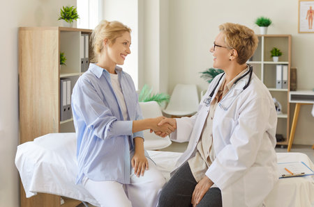 Handshake of doctor and young woman on medical consultation, patient and general practitioner shake hands with smiles. Girl showing trust, greeting professional physician in examination roomの写真素材