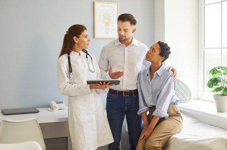 Consulting in clinic, female reproductologist doctor speaking holding tablet, helping couple, husband supporting wife listening information, professional and caring serious discussion, medical careの写真素材