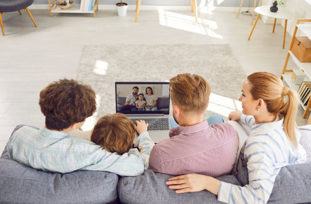 Back view of family with children sitting on sofa in living room, looking at screen, participating...