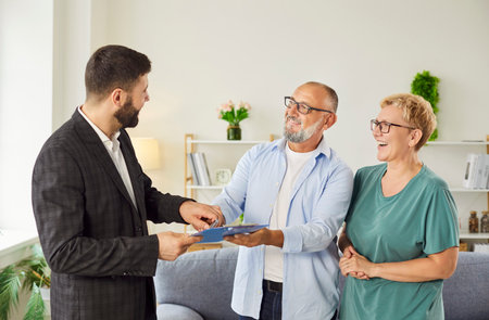 Real estate agent gives contract to smiling senior couple at home, explaining agreement and documents before signing to finalize the purchase and transfer. Realtor make a deal with new homeownersの写真素材