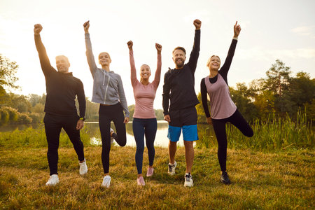 Group of happy fit sporty slim confident motivated diverse multiethnic athletes in sportswear standing on green river bank in park, raising hands up and smiling. Nature, sport, outdoor workout conceptの写真素材