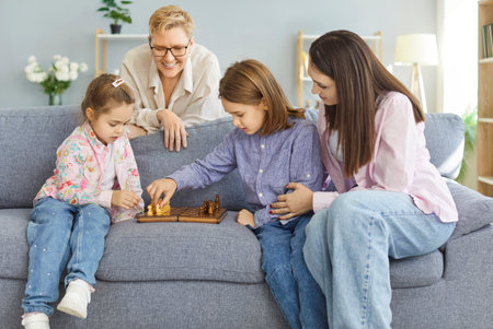 Two children of different age sitting on sofa, playing chess while two smiling adults watching and...