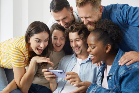 Man uses mobile phone to show vacation photos to his friends during meeting at home. Cheerful multiracial millennial people sitting together on sofa and smiling happily looking at smartphone screen.の写真素材