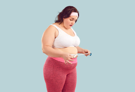 Young woman in sports bra and leggings looking at measuring tape that she holds around waist with surprised face expressionの写真素材