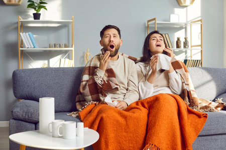 Young couple covered in blankets sitting on sofa at home with seasonal flu or cold. Sick unhappy man with woman sneezing suffering from runny nose and feeling unhealthy with influenza.の写真素材