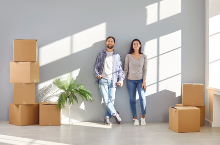 Happy young couple standing against the wall with a lot of cardboard boxes in a new apartment...