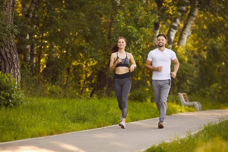 Smiling young man and woman jogging together along park path surrounded by green trees. Active couple enjoying morning run outdoors, maintaining healthy lifestyle, fitness routine and positive mood.の写真素材