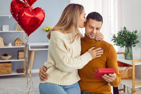 Happy loving couple with gift box hugging at home. Attractive husband holding present box and embracing wife who kissing him. Family couple celebrating Valentines Day with red heart shaped balloonsの写真素材