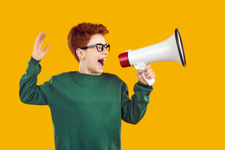 Funny red-haired handsome boy with glasses, dressed in green sweater, shouts with his mouth wide open into megaphone, pulling one hand back. Studio photo on isolated yellow background.の写真素材