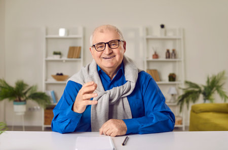 Smiling senior man having video call at home. Elderly man sitting at table, looking at camera and talking. Cheerful grandfather chatting with friends or family via video callの写真素材