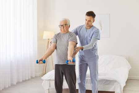 Health care worker helping senior elderly man to do exercises with dumbbells in nursing home....