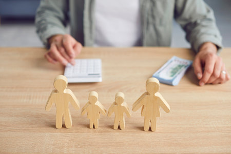 Soft focus closeup small wooden figures of mother, father and little children on table, with man managing family budget in blurred background. Finance, earning money, income conceptの写真素材