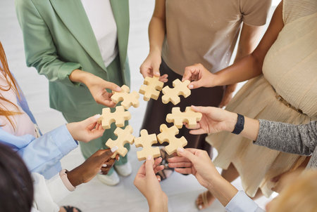 Teamwork hands hold puzzle pieces for solution. Colleagues link wooden jigsaw pieces in a circle, symbolizing collaboration, unity, and cooperation. Clear teamwork and partnership strategy.の写真素材