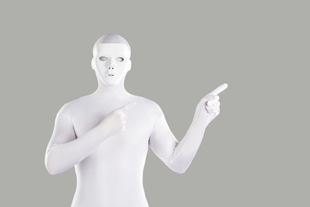 Mysterious anonymous man wearing white monochromatic mask and skintight nylon or spandex bodysuit costume standing on light grey background, and pointing his finger to sideの写真素材
