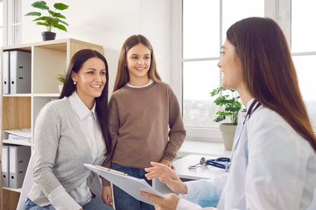Portrait of a cheerful teenager girl patient with her happy smiling mother talking with paediatrician doctor on consultation in clinic and listening the recommendations after medical examination.の写真素材