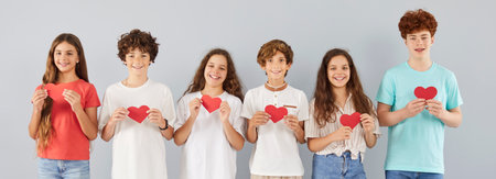Happy smiling group of young school children friends holding paper red hearts in hands together standing in a row and looking at camera isolated on gray background. Love and friendship concept.の写真素材