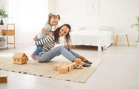 Mother and her little daughter share delightful moment together in their cozy living space. Surrounded by wooden toys, happy mother sits on soft rug, and her daughter playfully climbs on her back.の写真素材