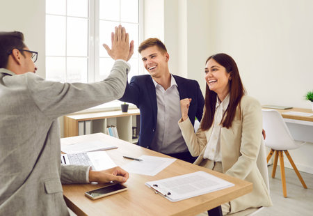 Happy joyful cheerful excited young couple celebrate success and give high five to loan broker, real estate agent or bank manager as they make business deal and sign contract during meeting in officeの写真素材